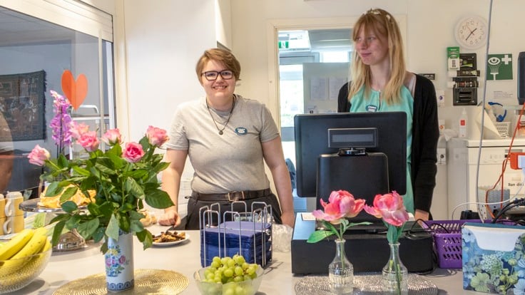 Två deltagare i cafélucka. Blommor och frukt på disken framför dem. 