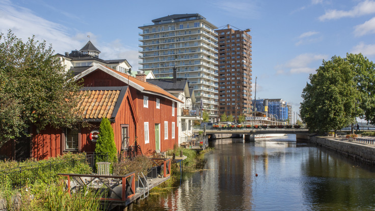 Rött trähus i förgrunden, vatten och två stora höghus