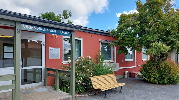 Putsad röd tegelbyggnad. En rosenbuske, ett  träd och  en träbänk syns framför bygganden.